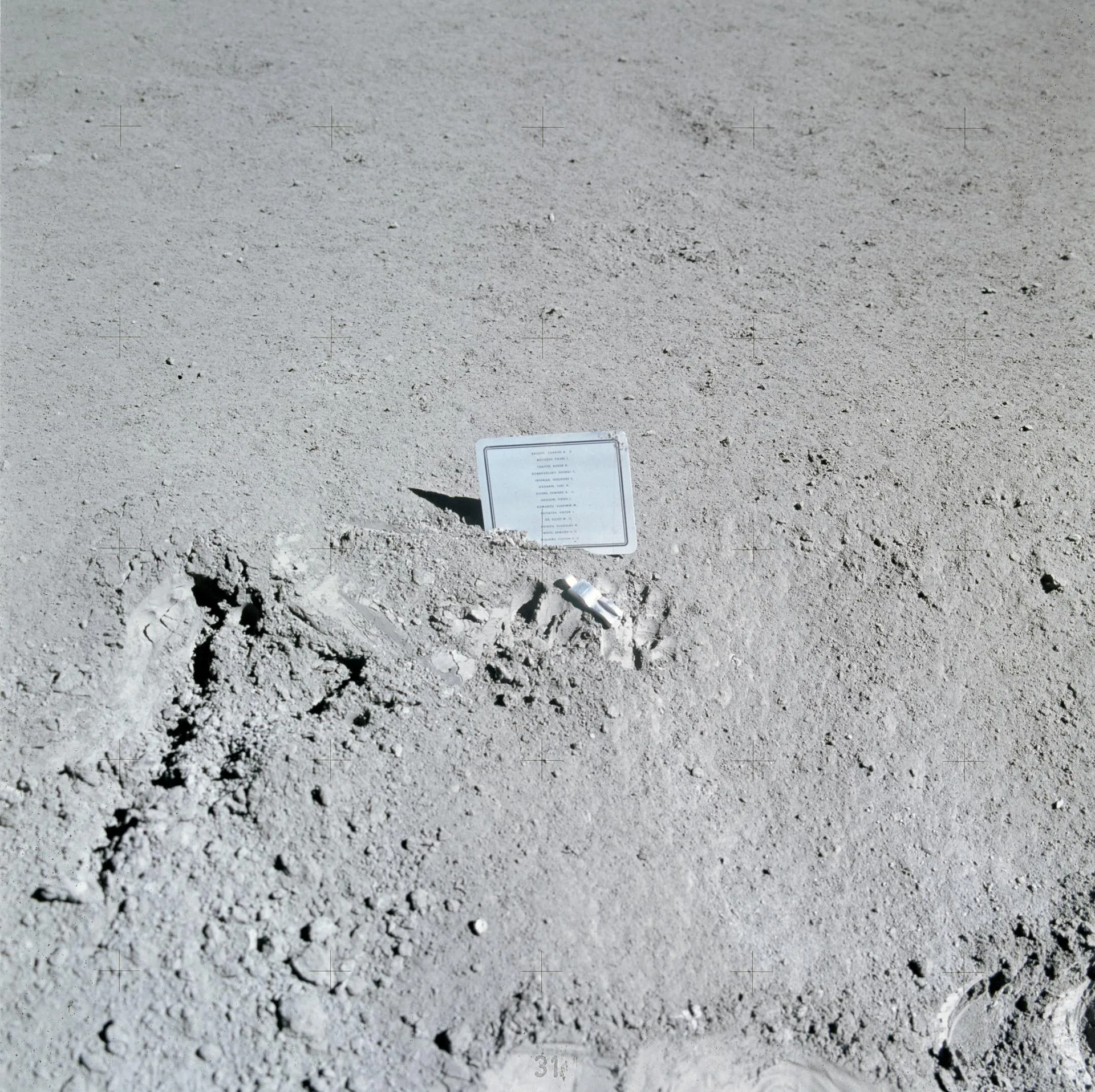 Apollo 15 ekibinin 1971'de Ay'daki Hadley-Apennine iniş bölgesine bıraktığı anma plaketi ve önünde yatan küçük "Düşen Astronot" heykelciği. Plakette hayatını kaybetmiş 14 astronot ve kozmonotun adı yazılı.